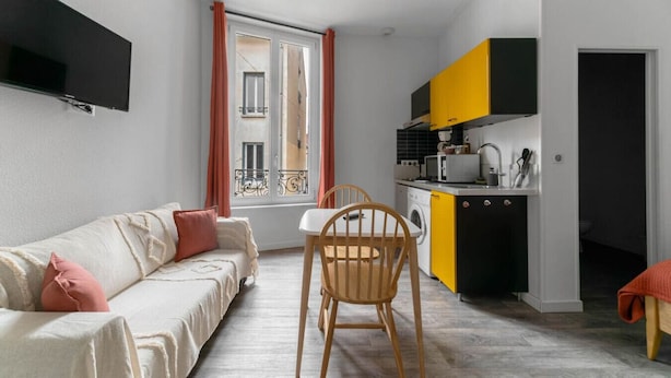 Een functionele leefruimte met een bank, eettafel, stoelen, een keuken met gele kastjes en een wasmachine.