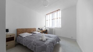 Une chambre à coucher avec un lit, des tables de chevet, une lampe, une fenêtre avec des rideaux et un radiateur.