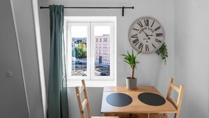 Une pièce avec une table en bois, deux chaises, une plante en pot, une horloge murale et une fenêtre donnant sur des immeubles.
