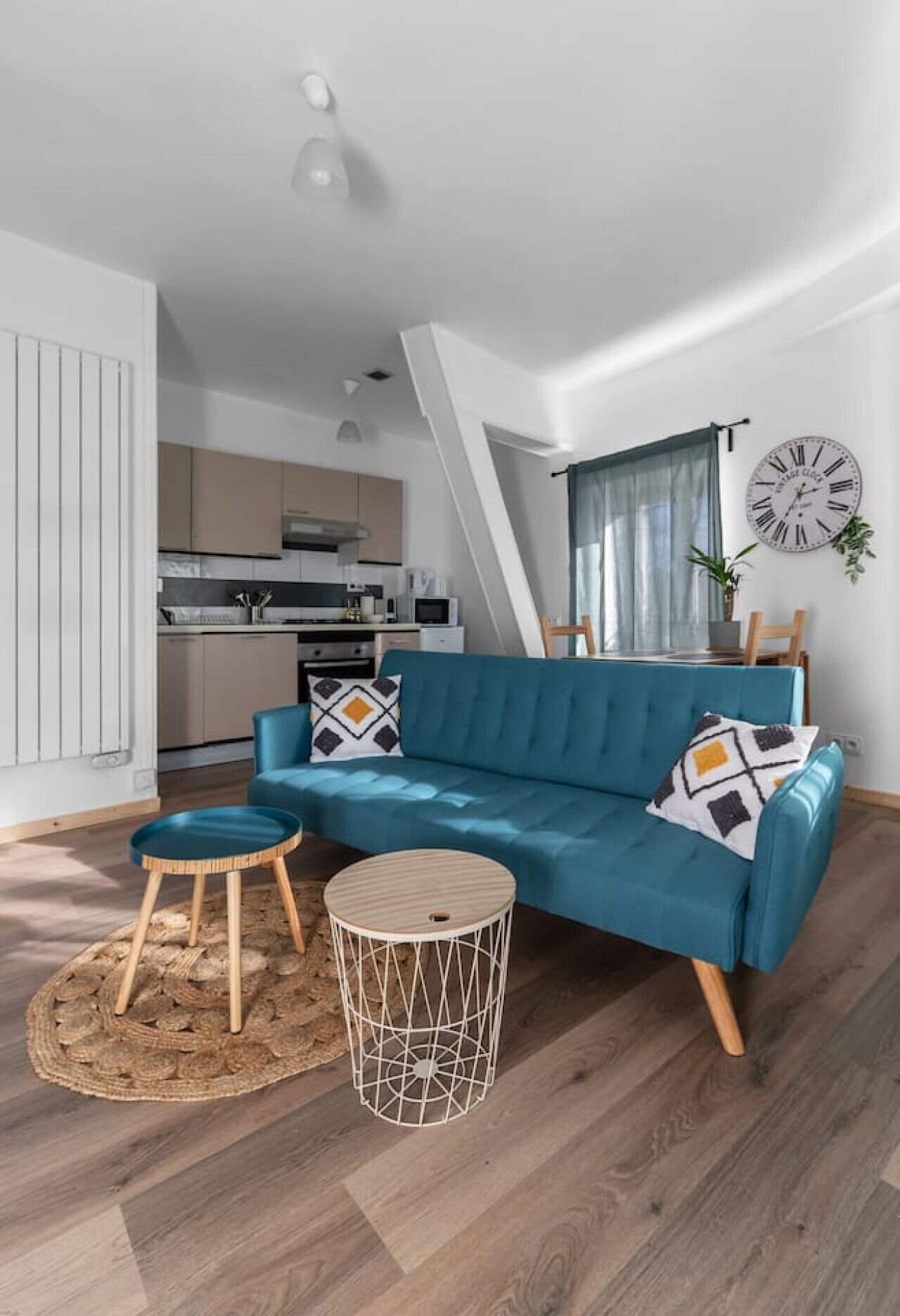 A modern living room with a blue sofa, two round tables, and a kitchen in the background.