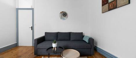 A living room with a dark grey sofa, a small round table with a plant, a round wooden stool, a white rug, and a wall-mounted decorative piece.