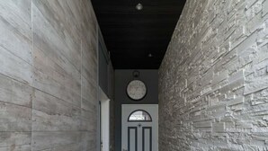 Un couloir avec un mur texturé d’un côté et un mur uni de l’autre, qui mène à une porte au-dessus de laquelle se trouve une horloge.