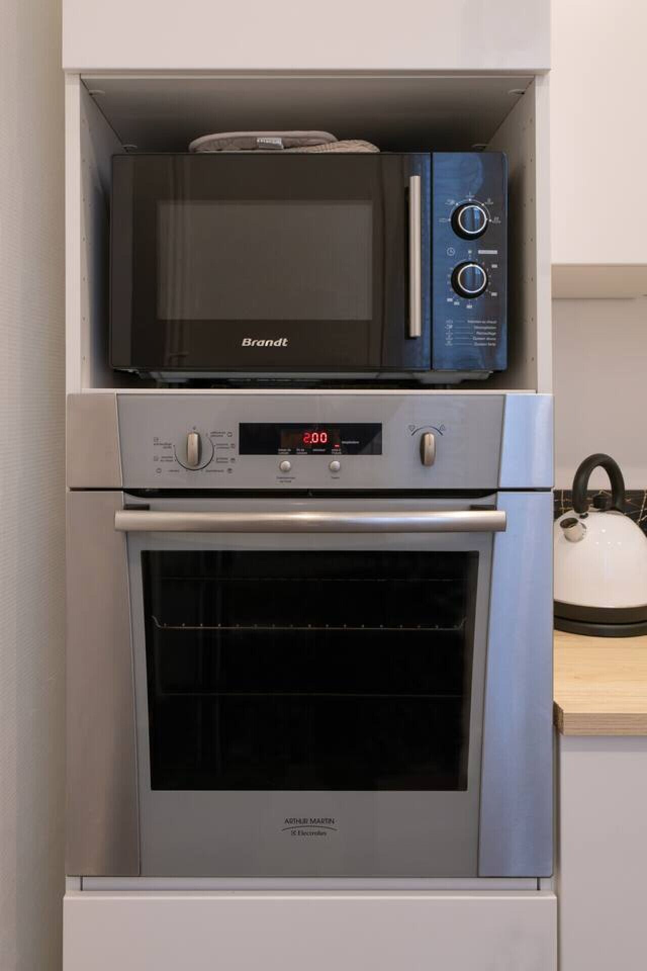Una cocina compacta con un microondas Brandt sobre un horno plateado, un reloj digital y una tetera en la encimera.