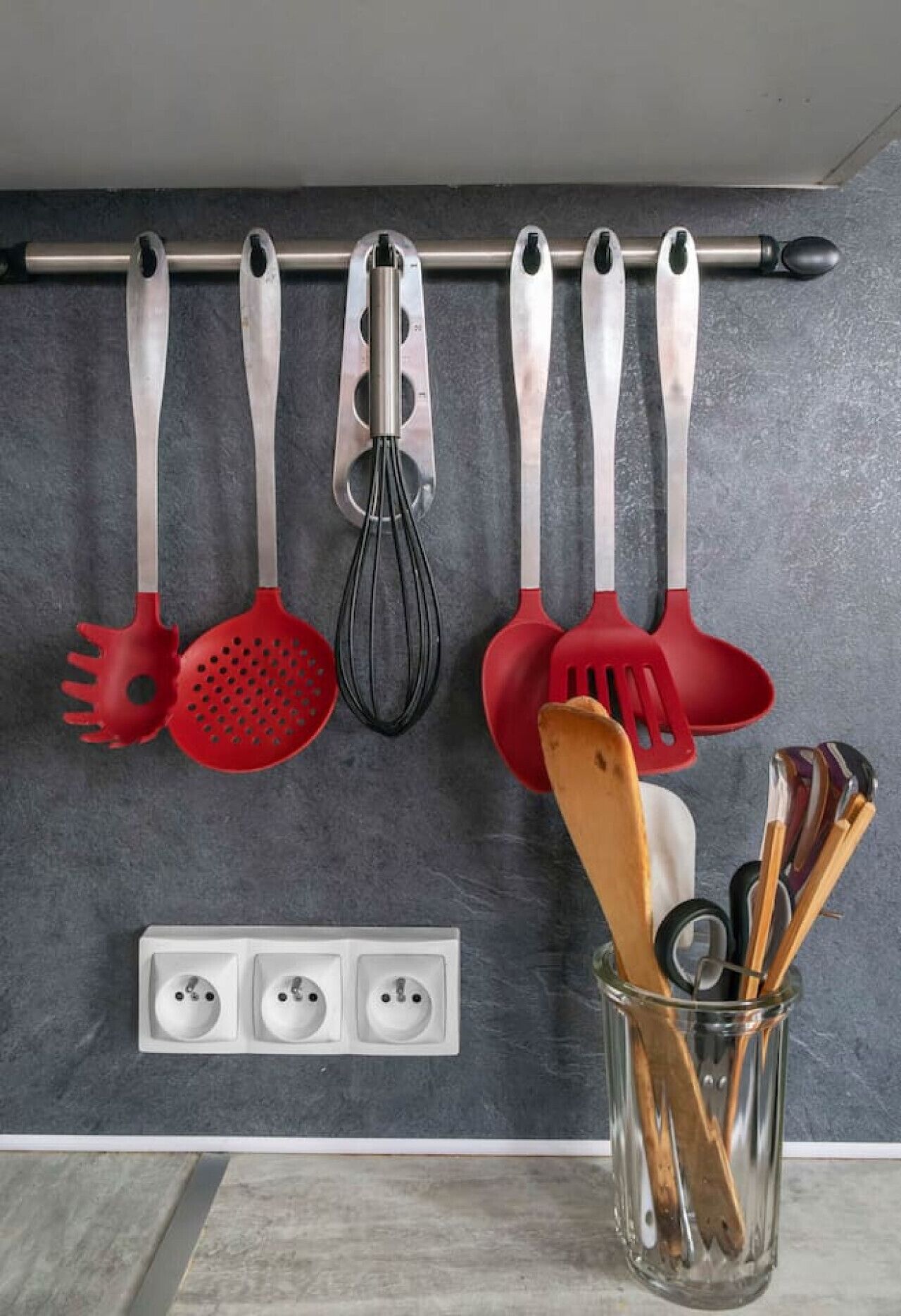 Een keuken met een messenrek vol keukengereedschap, een glazen houder met houten keukenspullen en een dubbel stopcontact aan de muur.