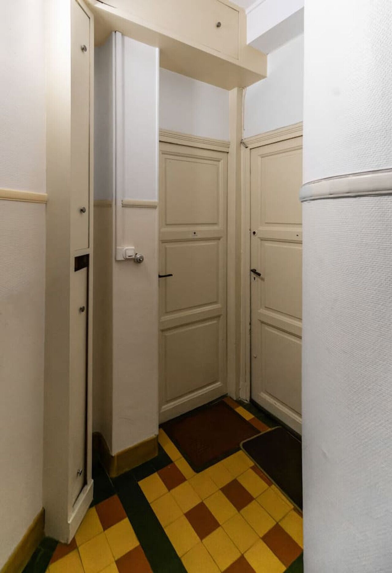 Un couloir étroit avec deux portes fermées et une porte entrouverte. Le sol est recouvert d’un motif à carreaux.
