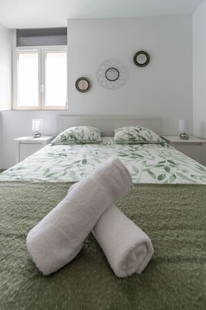 A neatly made bed with a green and white floral bedspread, two bedside tables with lamps, and a window with blinds. - Comfortable Air-cond apartment - city center (Vichy)