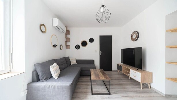 Ein modernes Wohnzimmer mit einem grauen Sofa, einem Holztisch, einem Fernseher auf einem Holzständer und einer geometrischen Pendelleuchte.