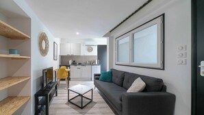 A modern living room with a sofa, coffee table, and TV stand. A kitchen is visible in the background with cabinets and a window. - Air-cond duplex apartment in the city center (Vichy)