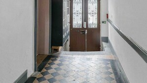 A hallway with patterned floor tiles, a wooden door with decorative glass panels, and a white wall with a small window. - Air-cond duplex apartment in the city center (Vichy)
