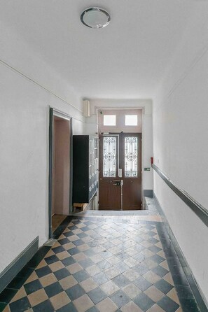 Leading to a door with a decorative grille, this hallway features a checkered floor and plain white walls.