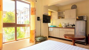 A compact hotel room with a bed, a small stool, a wooden table, a kitchenette with a microwave and refrigerator, and a window with curtains. - City View with Rooftop pool Studio (Nairobi)