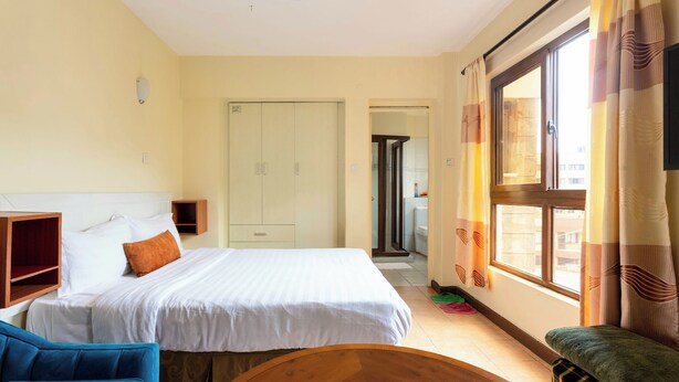 A hotel room with a bed, a blue chair, a wooden table, and a window with curtains. - City View with Rooftop pool Studio (Nairobi)