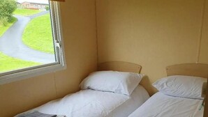 A small, single-room accommodation with two beds, a window with a view of a road and greenery, and a simple, functional design. - Lochside Retreat at Three Lochs Holiday Park (Newton Stewart)