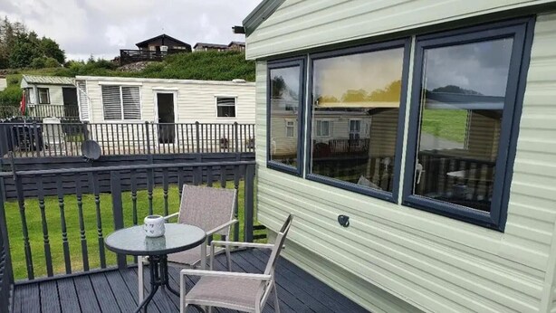 A deck with a table and chairs, a view of a house, and a grassy area. - Lochside Retreat at Three Lochs Holiday Park (Newton Stewart)