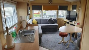 A compact RV interior with a kitchenette, a sofa, a small table, and a TV. - Lochside Retreat at Three Lochs Holiday Park (Newton Stewart)