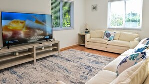 Ein Wohnzimmer mit einem Flachbildfernseher auf einem Holzständer, einem beigen Sofa mit gemusterten Kissen, einem blau-weißen Teppich und einem Fenster mit Jalousien.