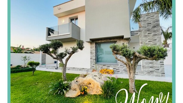 A modern villa with a well-maintained garden, a stone pathway, and a welcoming sign. - Modern Villa in Sicily, comfort, relax, garden and parking (Marsala)