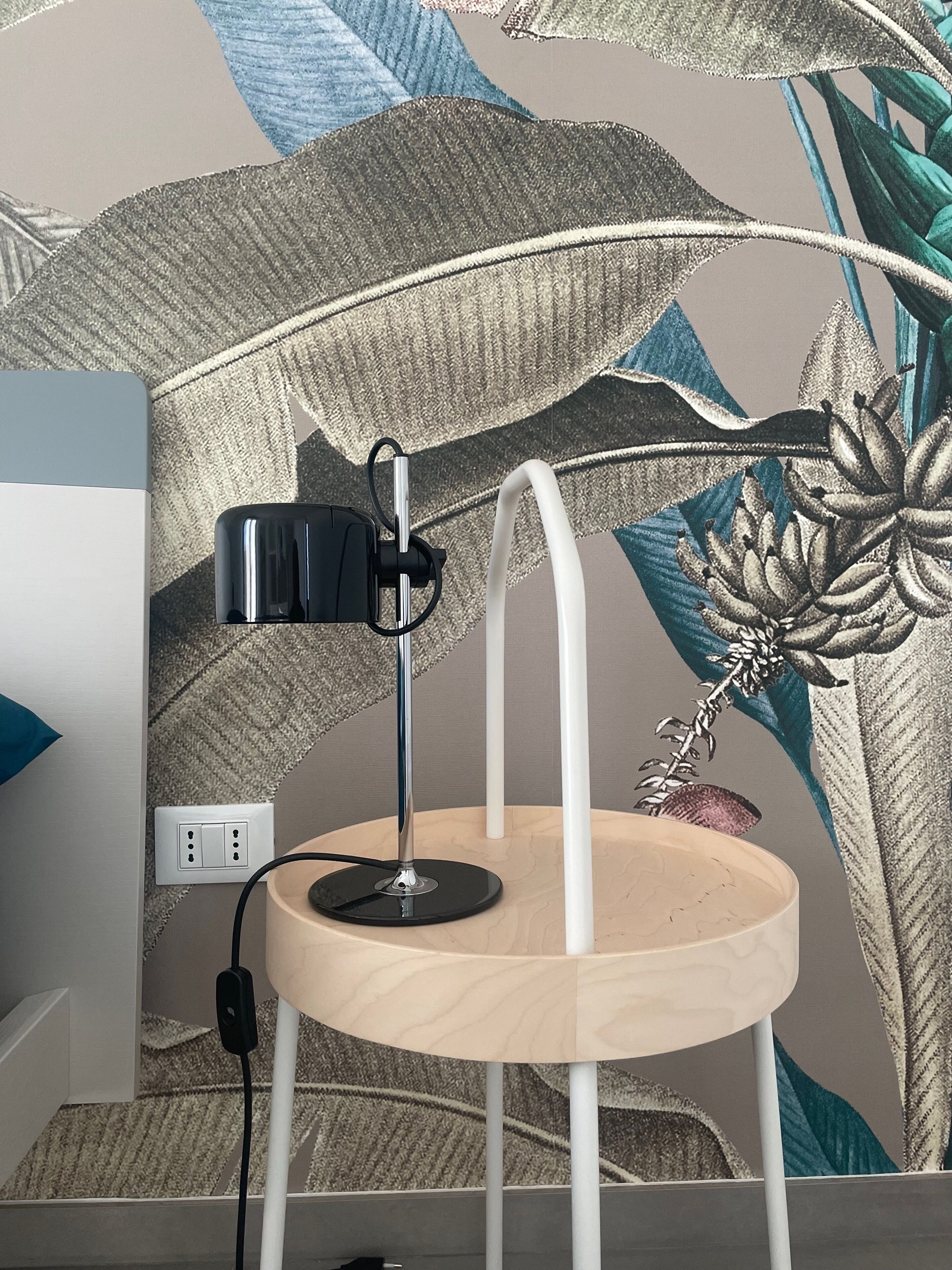 A desk lamp on a wooden stool with a power cord plugged into a wall socket.