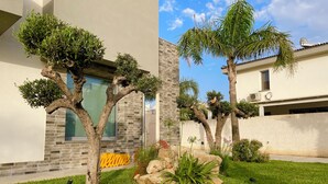 A well-maintained garden with various trees and shrubs, a clear sky, and a modern building with a brick facade. - Modern Villa in Sicily, comfort, relax, garden and parking (Marsala)