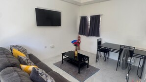 A modern living room with a sofa, a coffee table, a dining table with chairs, a ceiling fan, and a flat-screen TV mounted on the wall. - 1 Bed - Sleeps 4 - Parking - 5 Min to Beach (Ocho Rios)