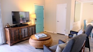 Ein Wohnzimmer mit Fernseher auf einer Holzkassette, einer blauen Tür und einem runden Ottomane aus Weidengeflecht.