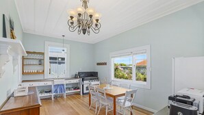 Une cuisine avec des meubles blancs, une table à manger avec des chaises, un lustre et une vue sur l’extérieur grâce à de grandes fenêtres.