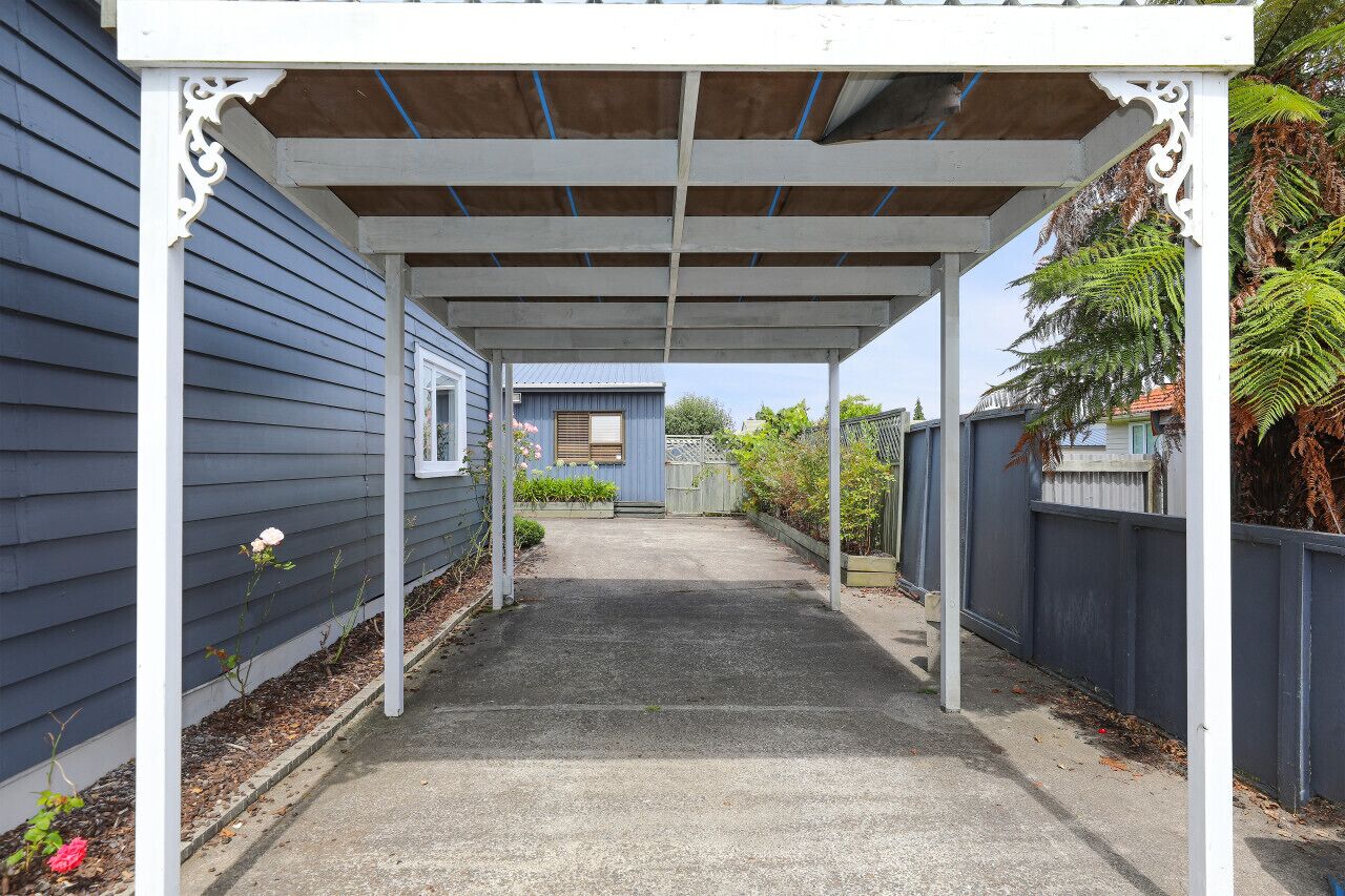 A covered outdoor area with a concrete pathway, white columns, and a blue house in the background.