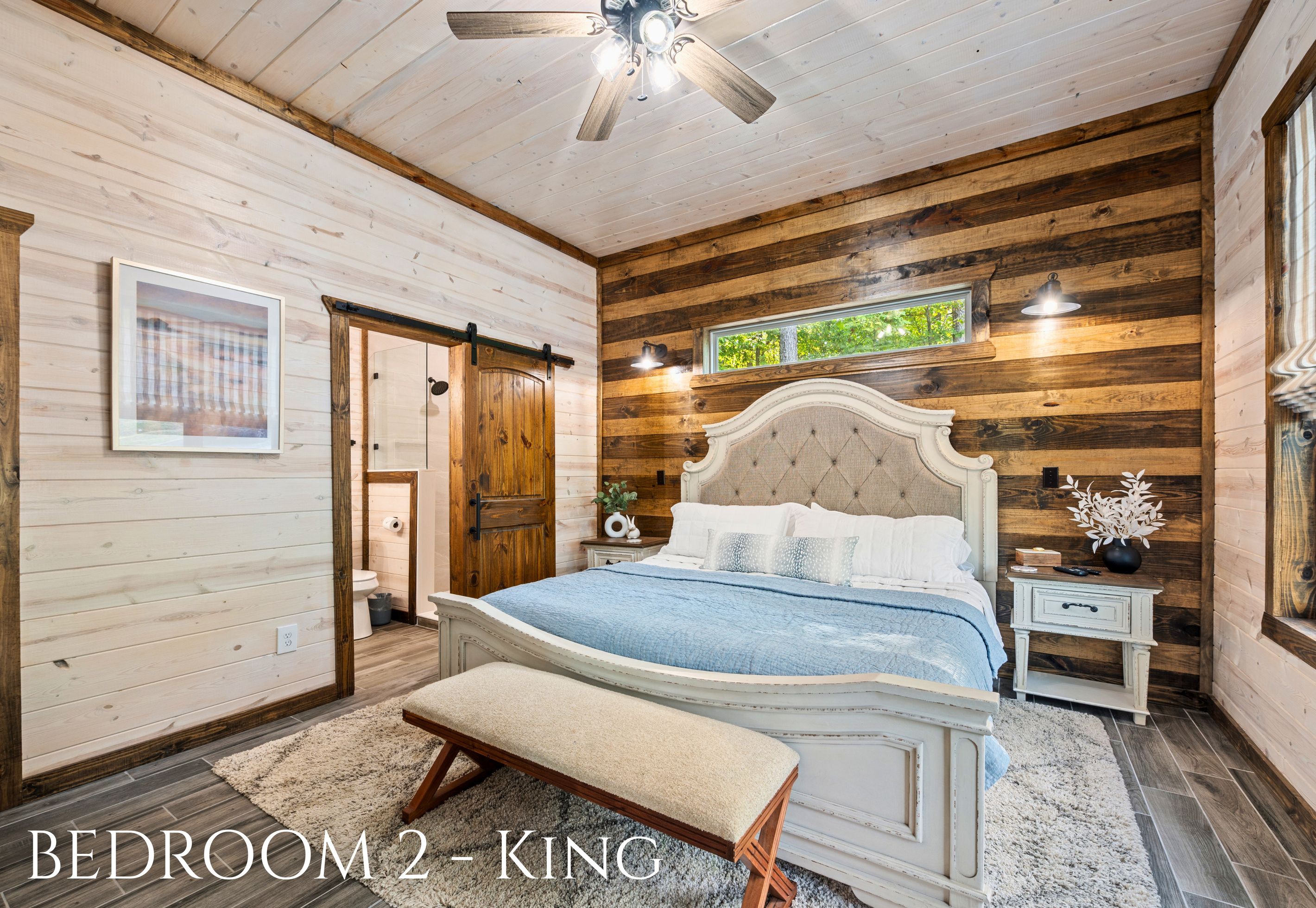 A bedroom with a wooden log wall, a bed with a blue bedspread, a bench, a nightstand, and a ceiling fan.