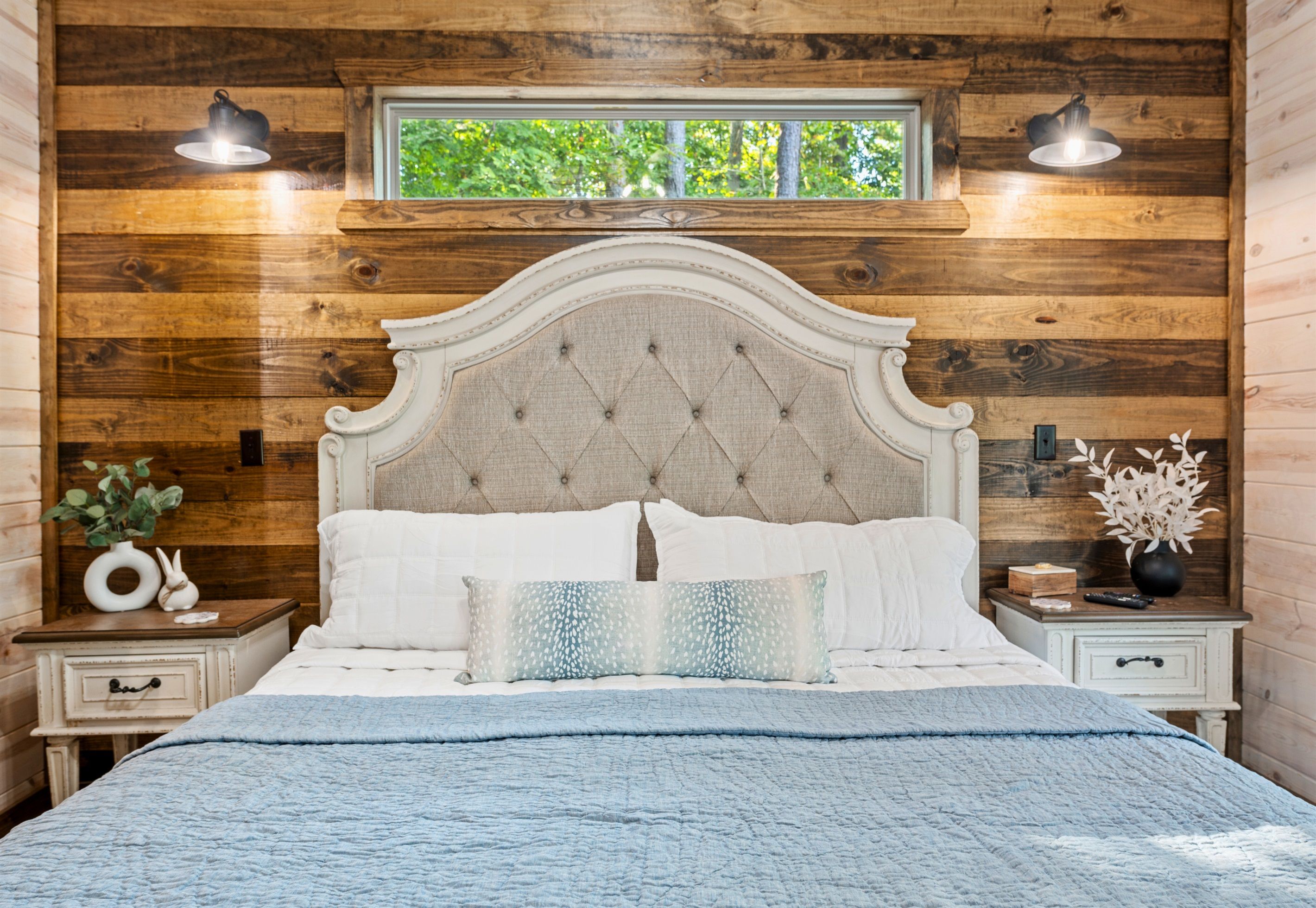 Een slaapkamer met een houten wandpaneel, een gestoffeerd hoofdbord, een raam, twee bedlampen, een nachtkastje met een vaas, en een bed met een blauwe deken.