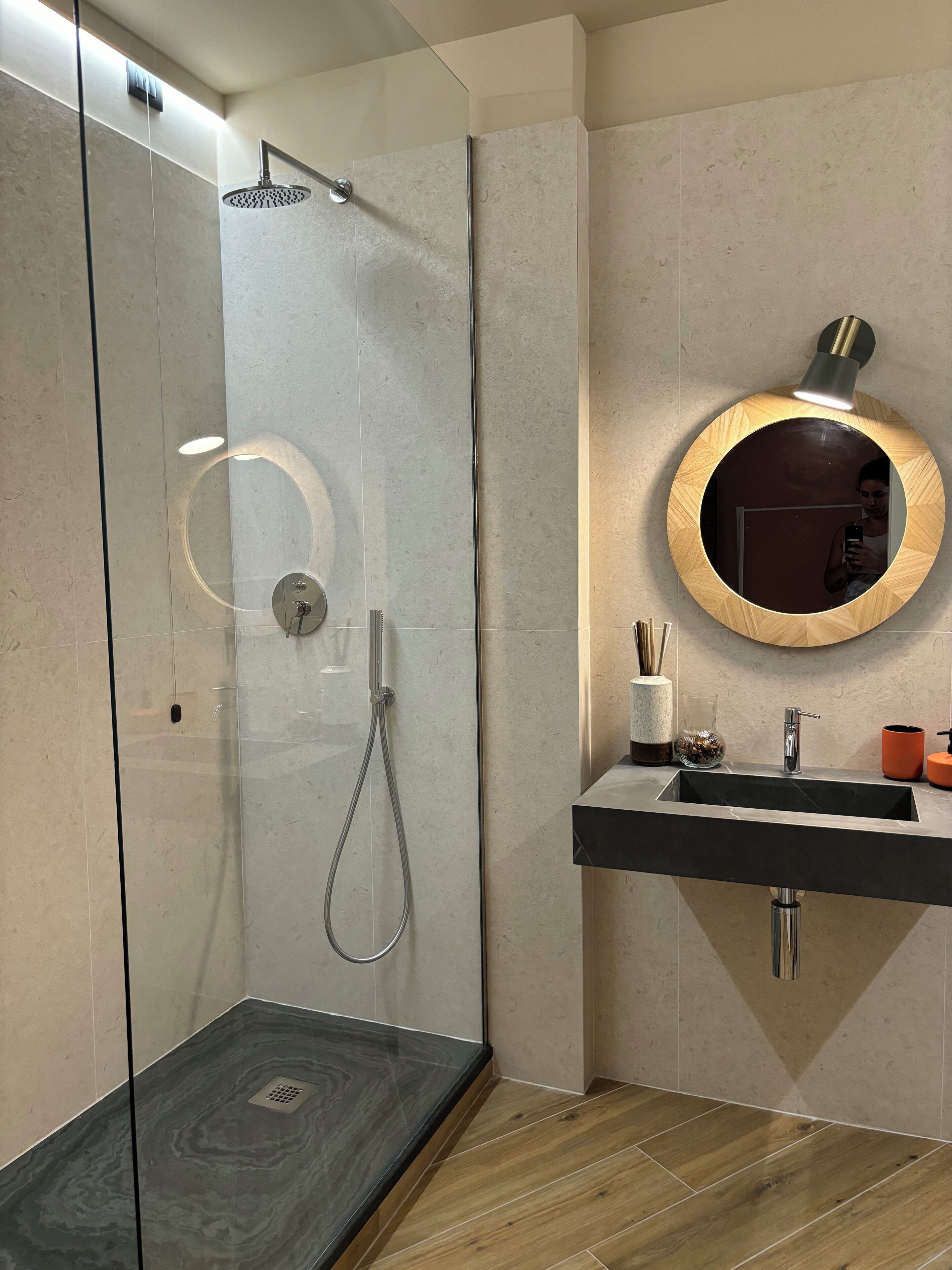 A modern bathroom with a glass-enclosed shower, a round mirror with a wooden frame, and a black sink with a chrome faucet.
