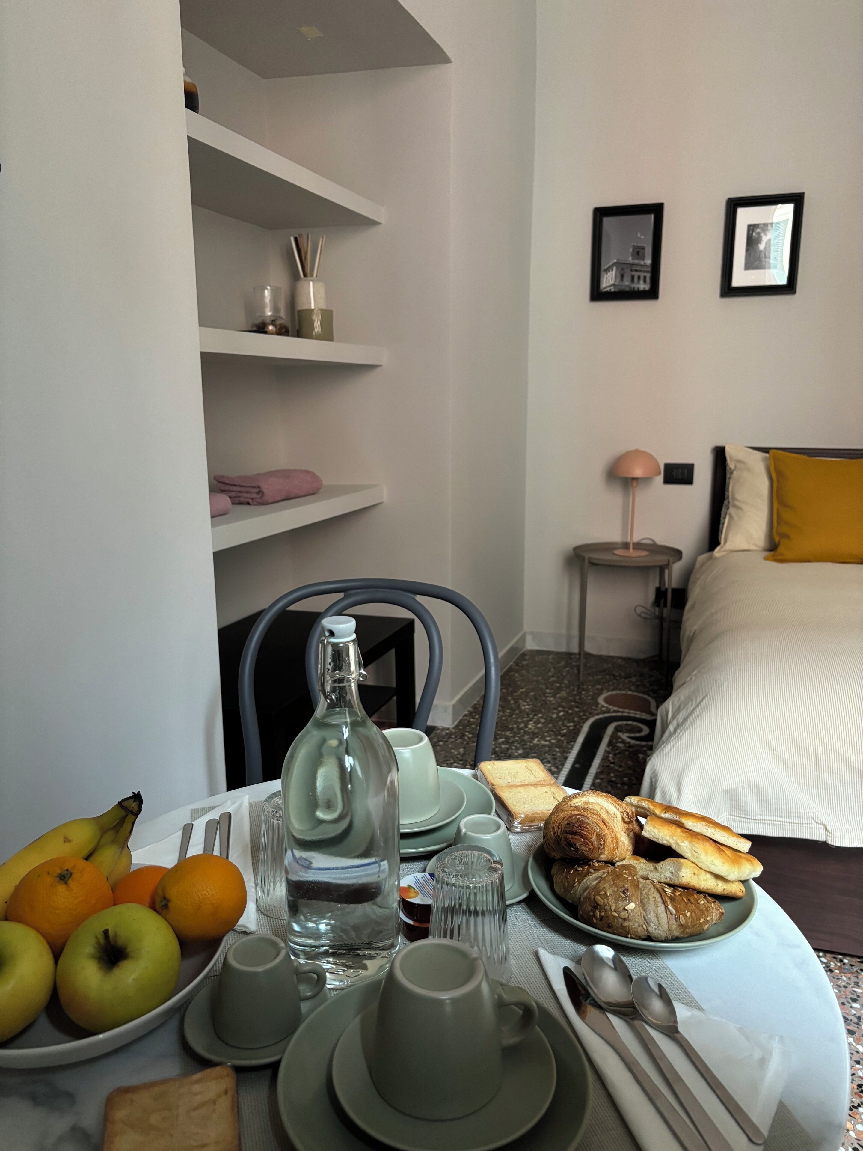 A neatly arranged breakfast table with fruits, bread, and a bottle of water, a bed with a yellow pillow, and a nightstand with a lamp in a hotel room.