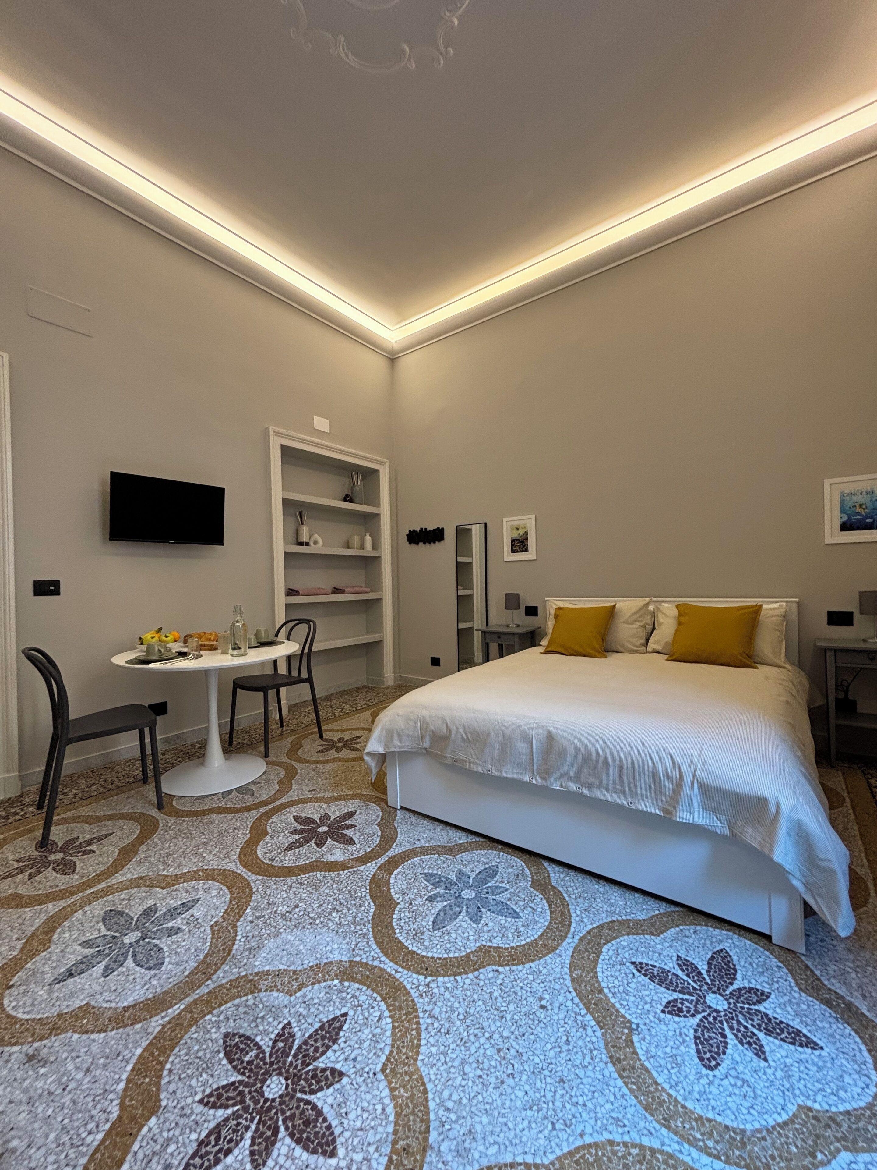 A bedroom with a bed, a desk, and a chair. There is a television on the wall and a shelf with decorative items.