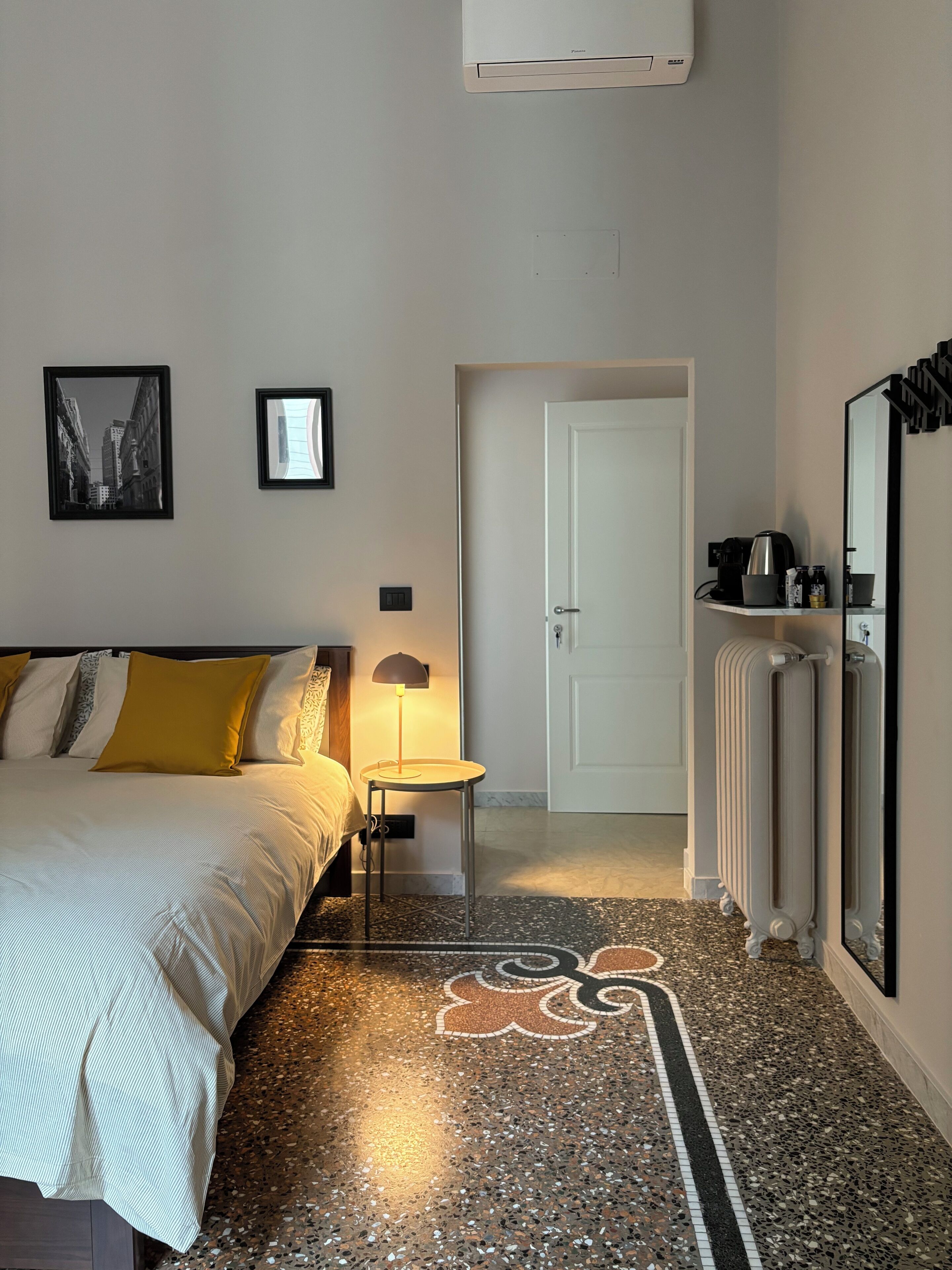 A hotel room with a bed, a small table with a lamp, a radiator, and a door leading to another room.
