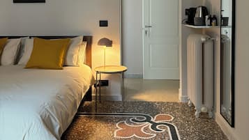 Habitación de hotel con cama, una mesita con lámpara, un radiador y una puerta que da a otra habitación.