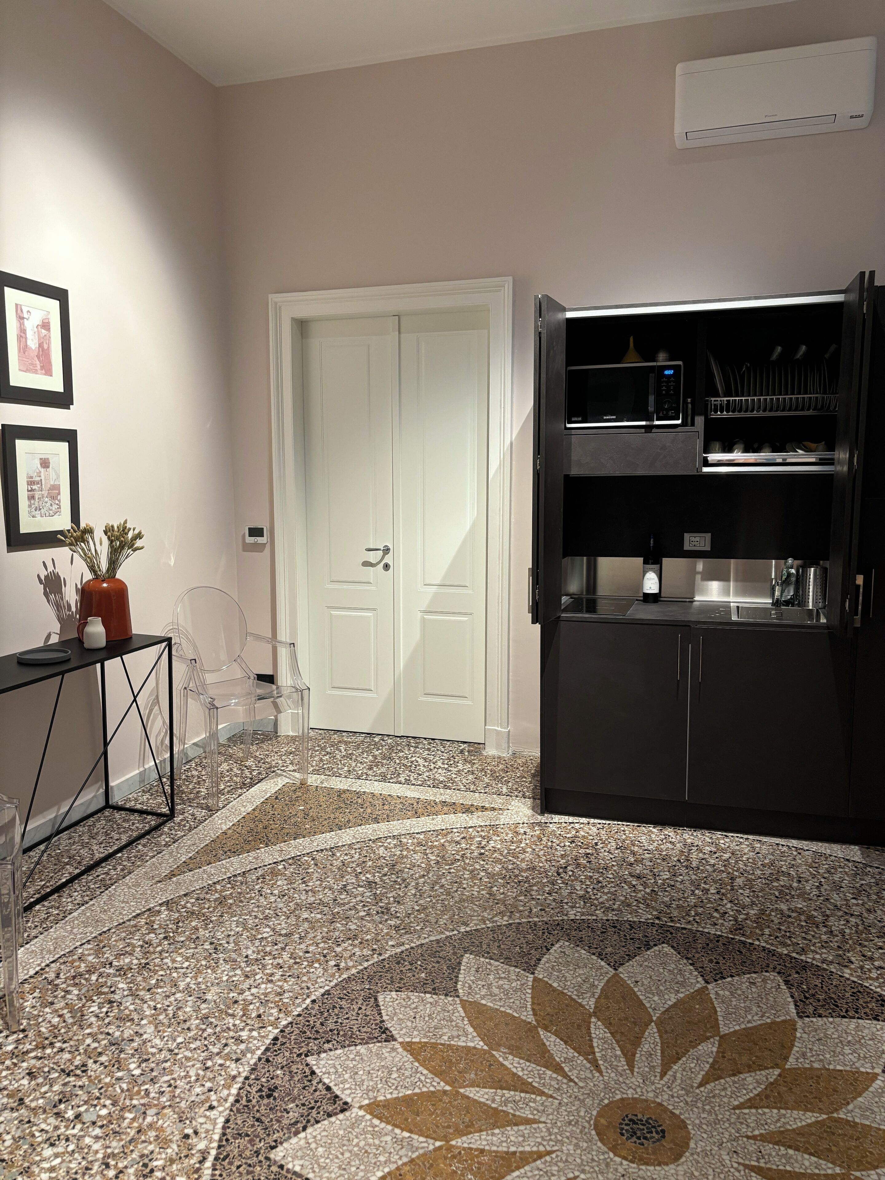 A modern kitchen with a microwave, a coffee machine, and a built-in oven. There is a door with a number on it, a small table with a vase and a cup, and a patterned floor.