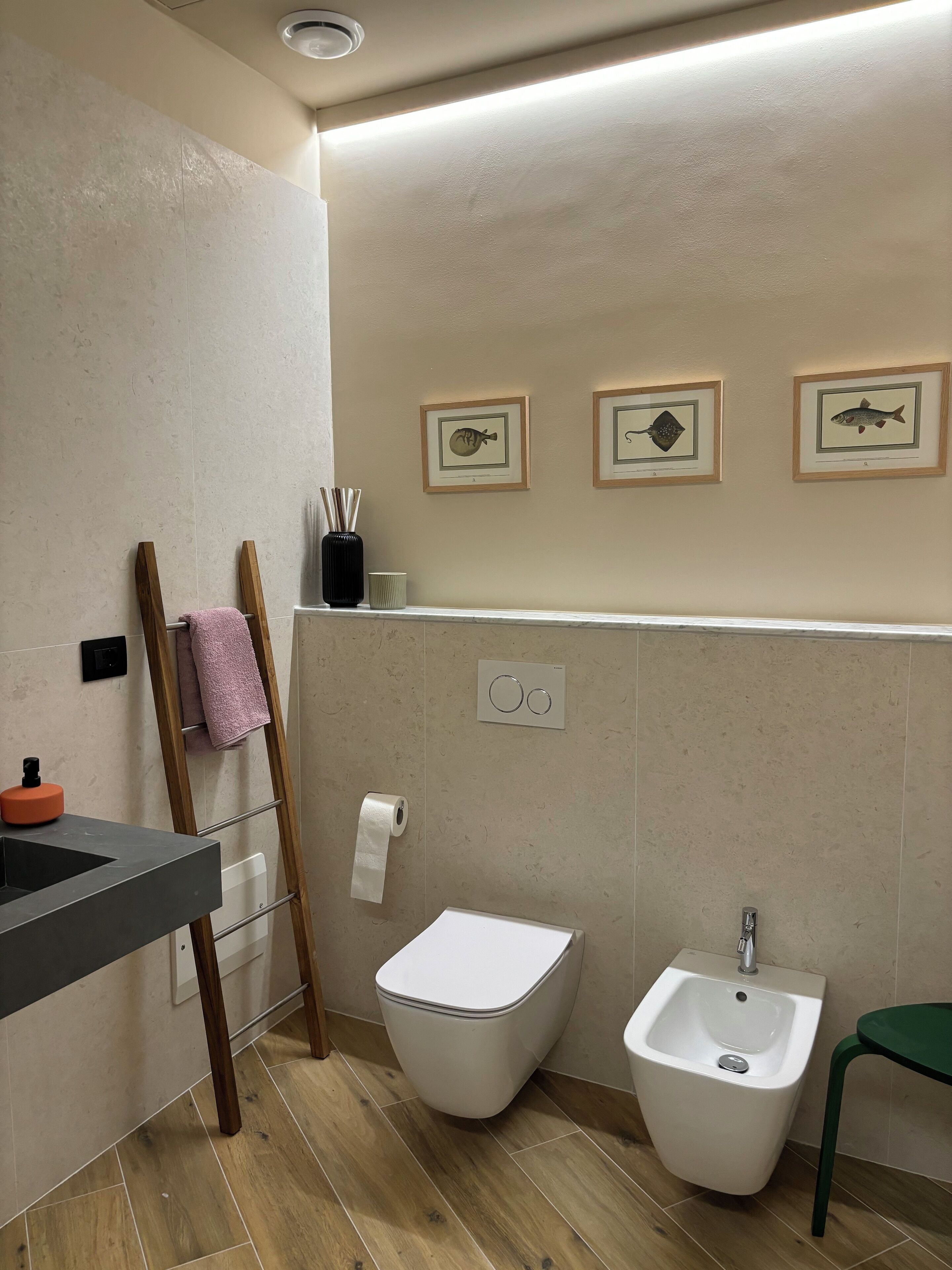 A modern bathroom with a toilet, bidet, and sink. There are framed pictures on the wall and a towel rack with a pink towel.