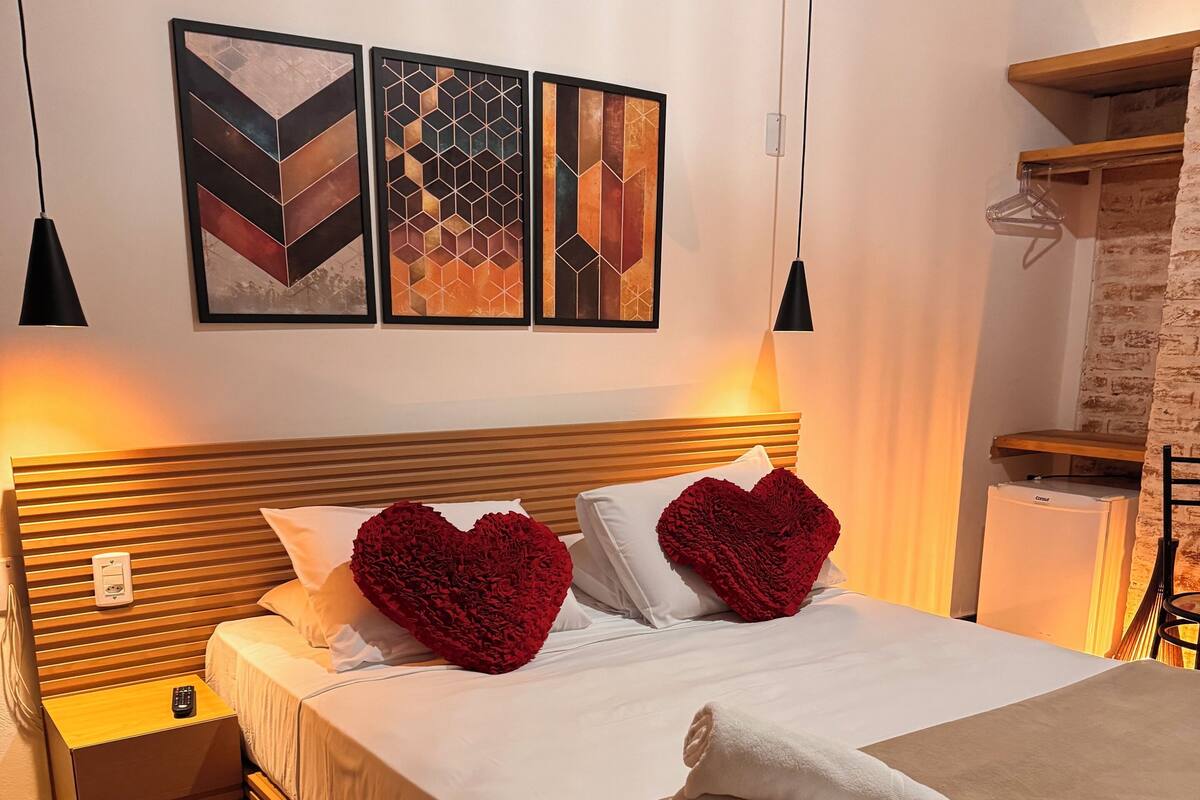 Une chambre d’hôtel avec un lit, des oreillers en forme de cœur rouge, une couvre-lit marron et un tapis rouge. Deux œuvres d’art sont accrochées au mur au-dessus du lit, et une petite table de chevet est placée à côté, avec une télécommande dessus.