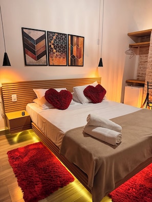 A hotel room with a bed, red heart-shaped pillows, a brown bedspread, and a red rug. There are two wall-mounted artworks above the bed and a small nightstand with a remote control. - Pousada Bosque (Camanducaia)