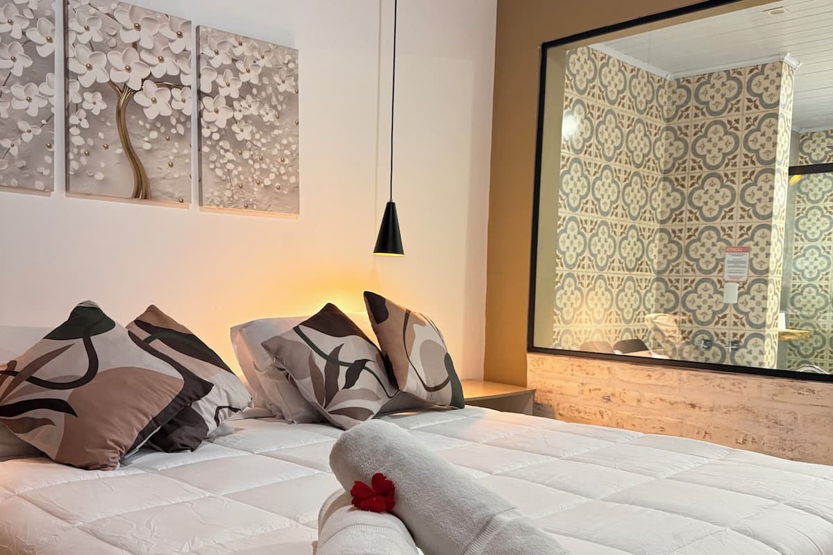 Un lit bien fait, avec une serviette enroulée et une fleur rouge, un luminaire suspendu noir et un grand miroir qui reflète un papier peint à motifs.