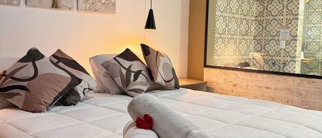 A neatly made bed with a rolled towel and a red flower, a black pendant light, and a large mirror reflecting patterned wallpaper.