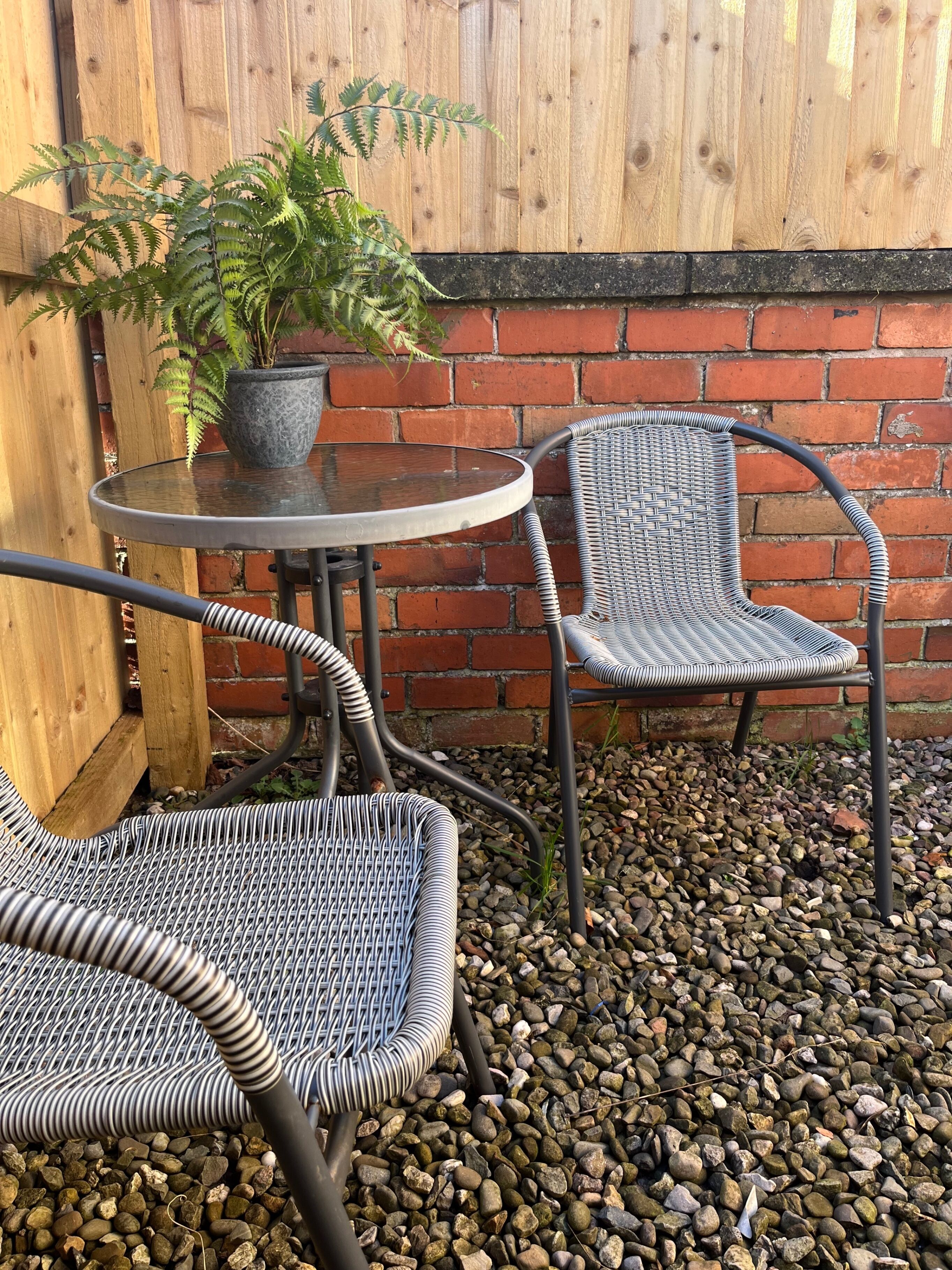 Un espace patio avec une table ronde et deux chaises en osier, une plante en pot et un mur en briques.