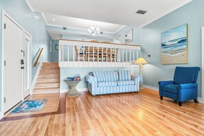 A spacious living room with a wooden staircase, a blue sofa, a blue armchair, and a beach-themed painting on the wall.