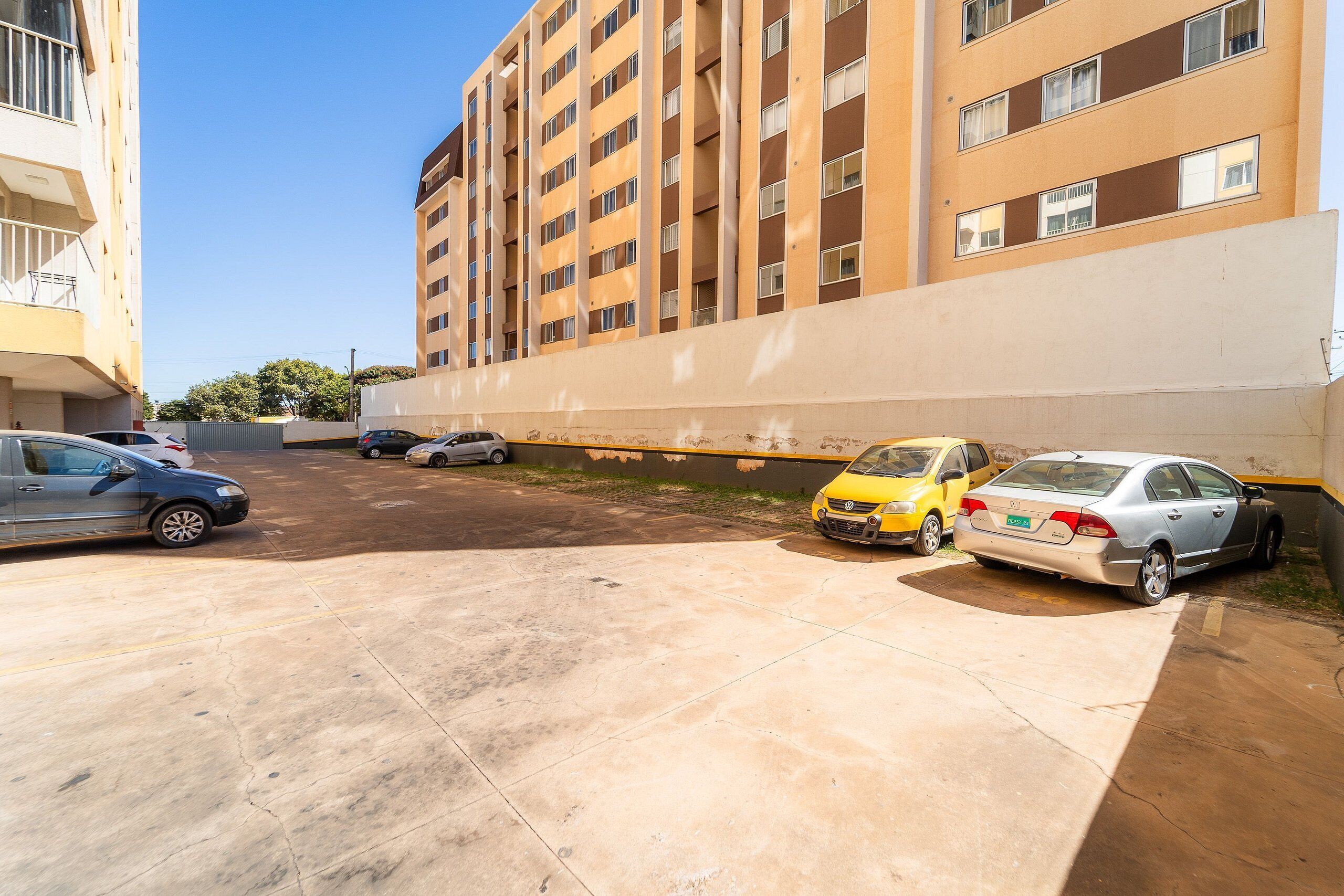Uma área de estacionamento com vários carros, um edifício de fachada bege e céu claro.