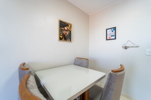 A dining area with a white table and chairs, a framed artwork on the wall, and a small shelf with a sign.