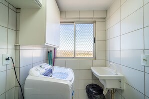 A small, compact bathroom with white tiles, a window with a metal grid, a washing machine, a sink, and a trash bin.