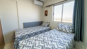A room with two beds, a window with a view, an air conditioning unit, and a door.