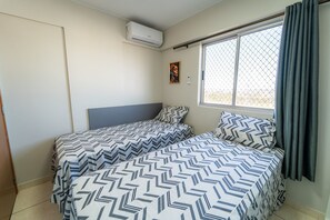 A room with two beds, a window with a view, an air conditioning unit, and a door.