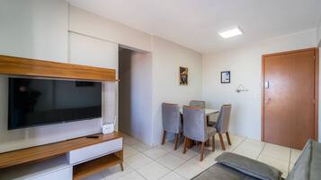 Uma sala de estar com televisão de tela plana, um móvel para aparelhos eletrônicos de madeira, uma mesa de jantar com cadeiras, um sofá e uma porta.