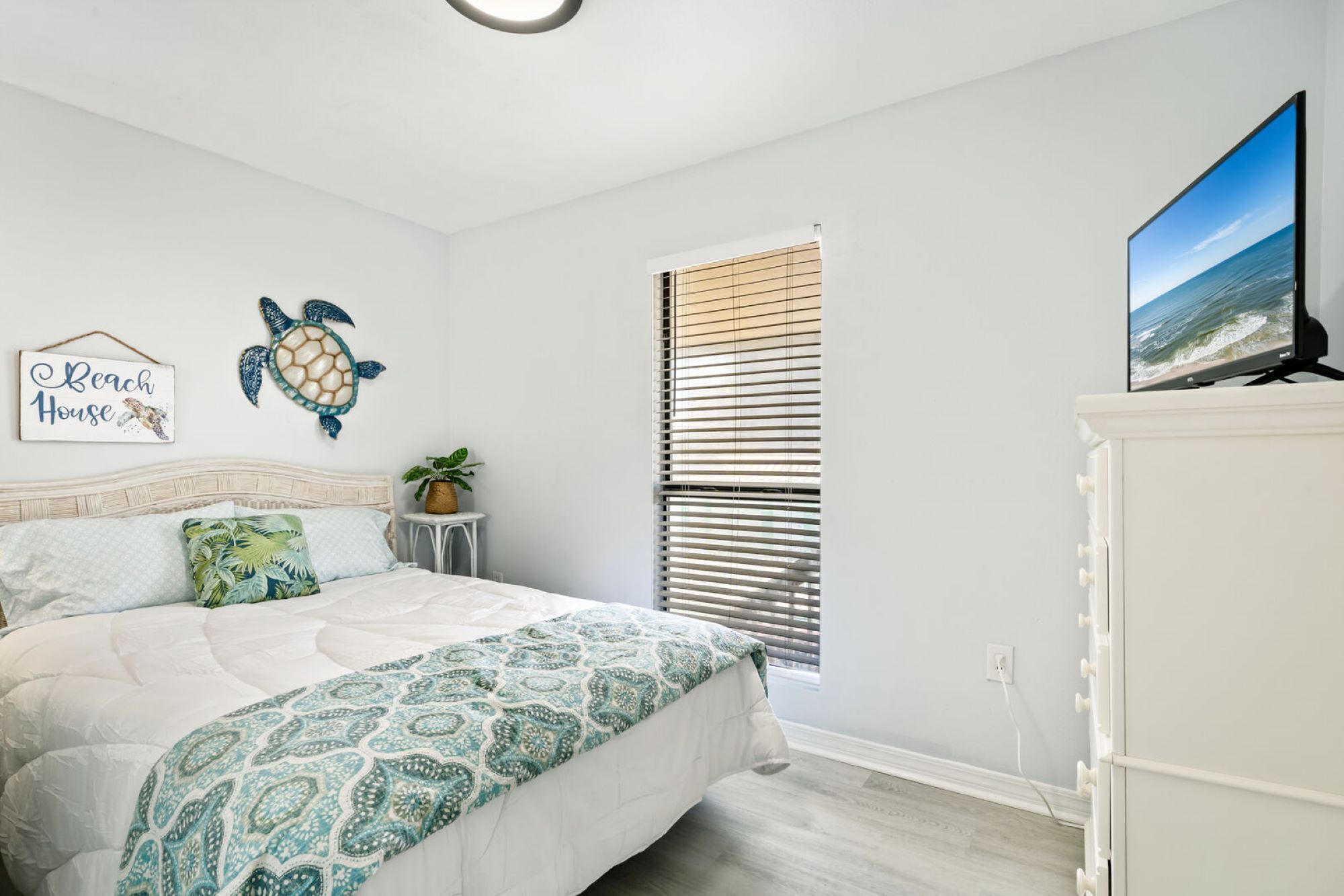 Un dormitorio con cama, televisión instalada en la pared, ventana con persianas, una mesita con una planta y un cartel decorativo.