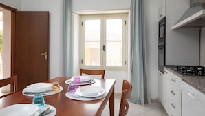 A dining area with a wooden table set for four, a window with curtains, and a kitchen with white cabinets and a stove. - Casa Bata by Wonderful Italy (Budoni)